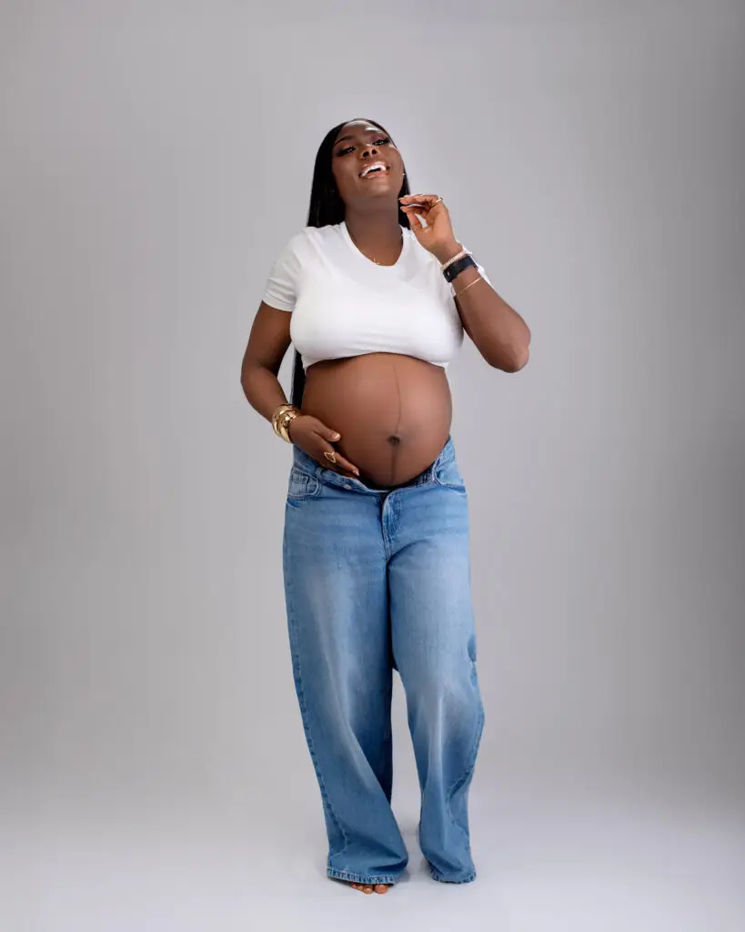 Pregnant woman in a white shirt and oversized jeans joyfully poses, showcasing her baby bump against a neutral background.