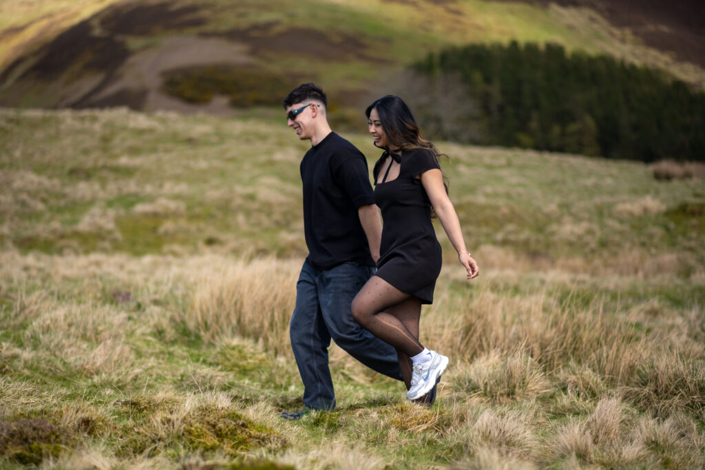 A couple joyfully walks hand in hand through a grassy landscape, enjoying a sunny day outdoors.
