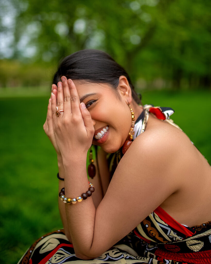 A smiling woman playfully covers her face with one hand while sitting in a lush green park.