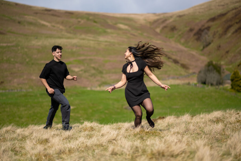 A man and woman joyfully dance on a grassy hillside, surrounded by rolling green hills under a bright sky.