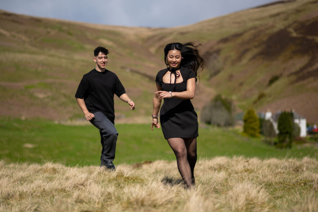 Two friends joyfully run through a grassy field with rolling hills in the background on a sunny day.
