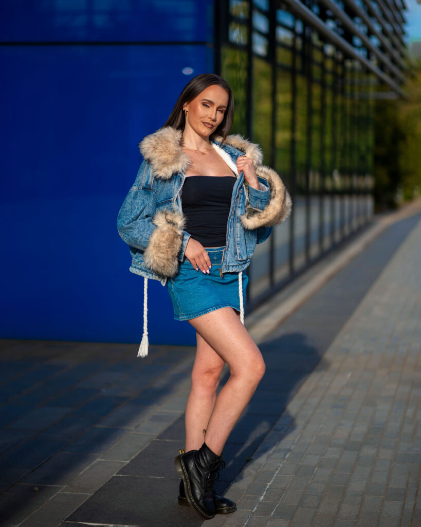 A woman poses confidently in a stylish denim jacket with fur trim, black top, and denim skirt against a blue background.