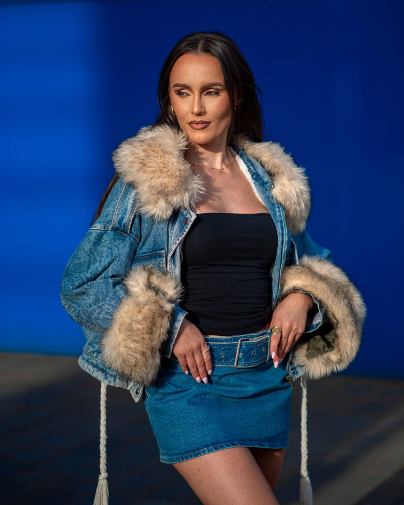 A woman poses confidently in a stylish denim jacket with fur trim and a matching denim skirt against a vibrant blue background.