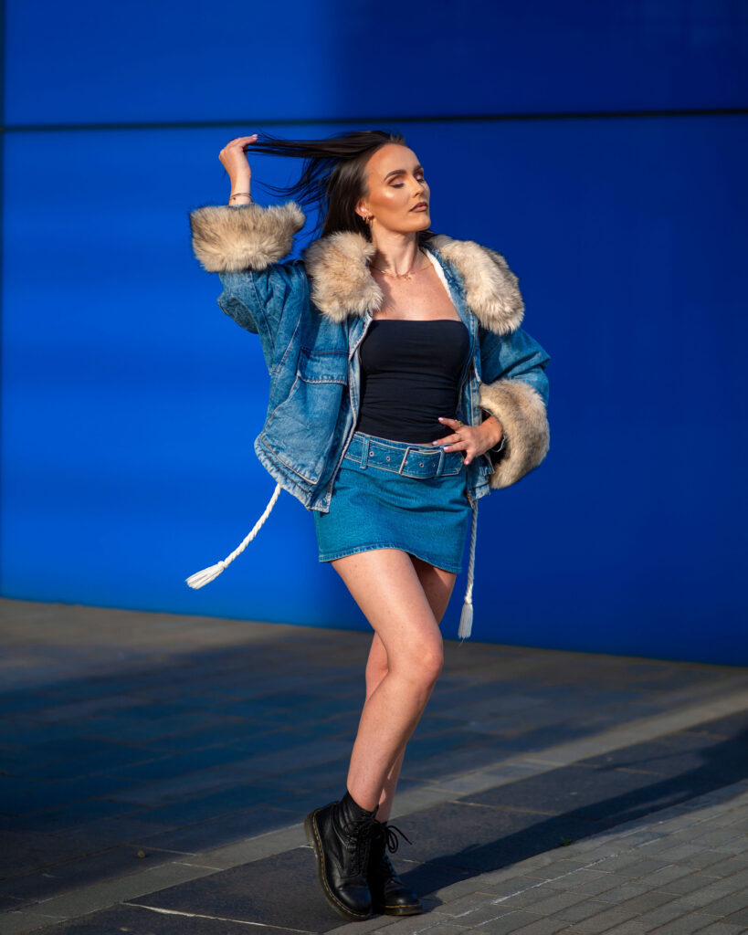 A woman poses confidently in a stylish denim outfit with a fur-lined jacket against a vibrant blue background.
