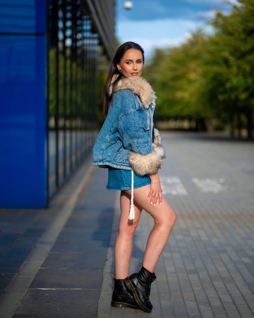 Model poses confidently in a stylish denim jacket with fur trim, showcasing a trendy outfit against a modern urban backdrop.
