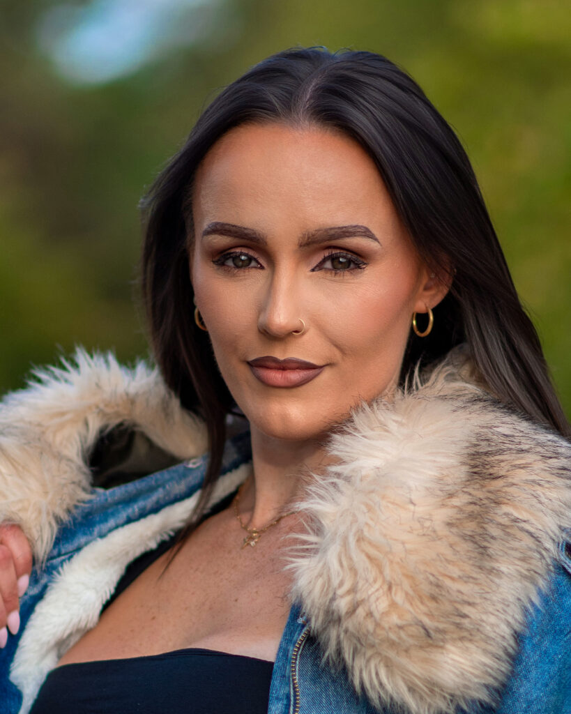 Woman with long dark hair and a fur-lined denim jacket poses confidently outdoors, showcasing her makeup and style.